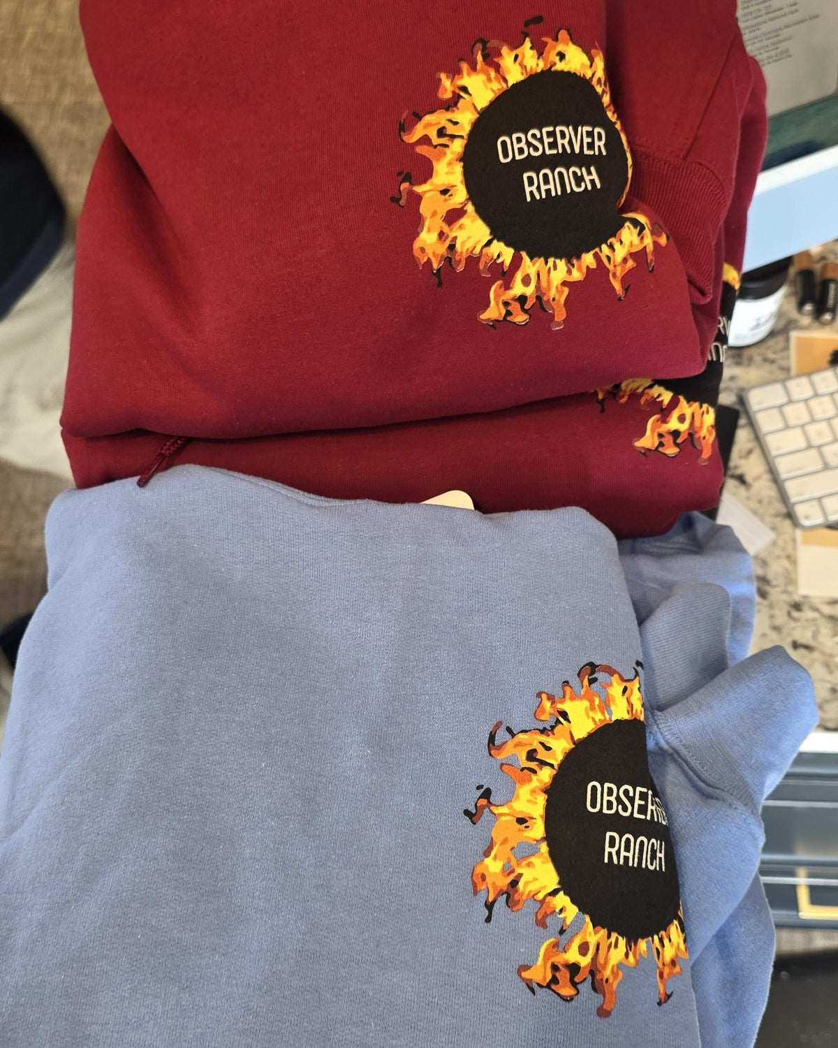 Two stacked thick pullover hooded sweatshirts with the Observer Ranch flaming sun logo on the chest, cardinal red on top and light blue below