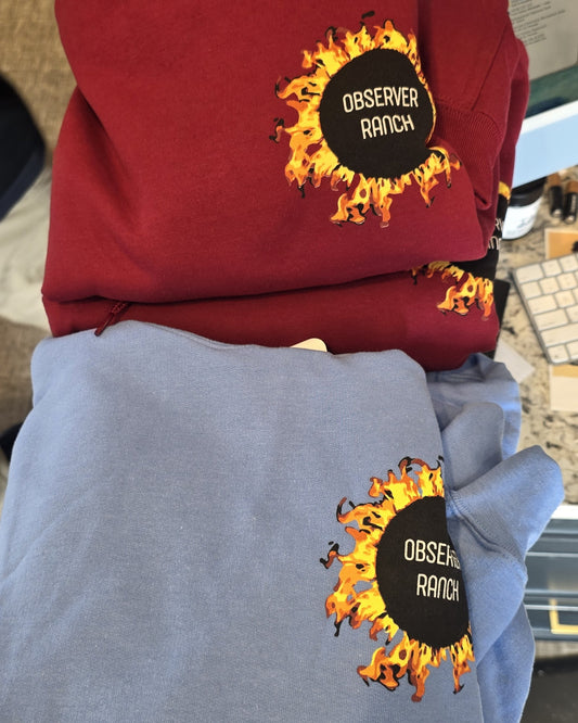 Two stacked thick pullover hooded sweatshirts with the Observer Ranch flaming sun logo on the chest, cardinal red on top and light blue below